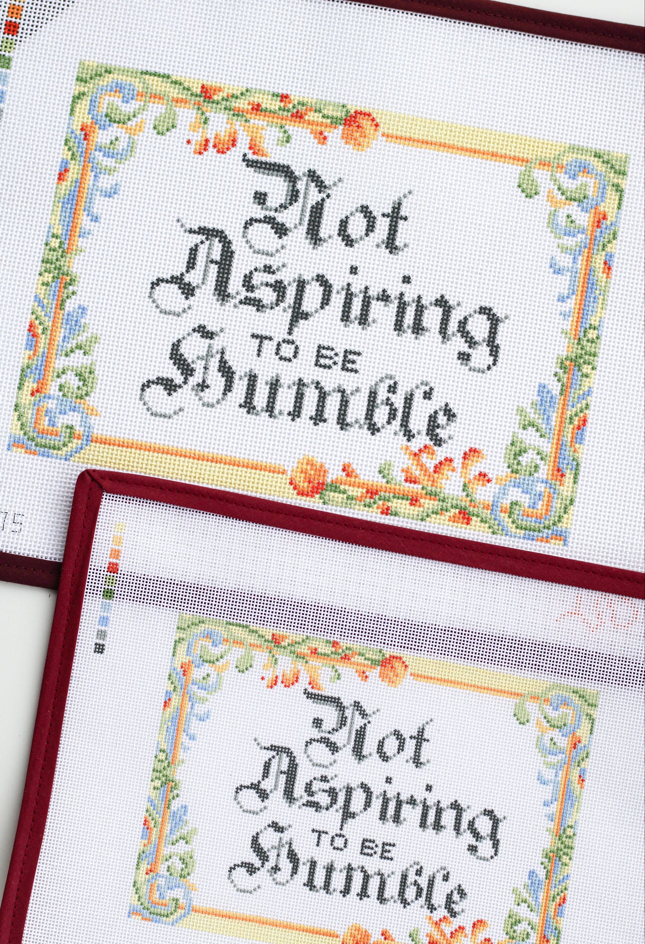 Two layered views of the Not Aspiring to Be Humble needlepoint canvas showcasing the medieval-inspired floral border and gothic lettering in different lighting angles.