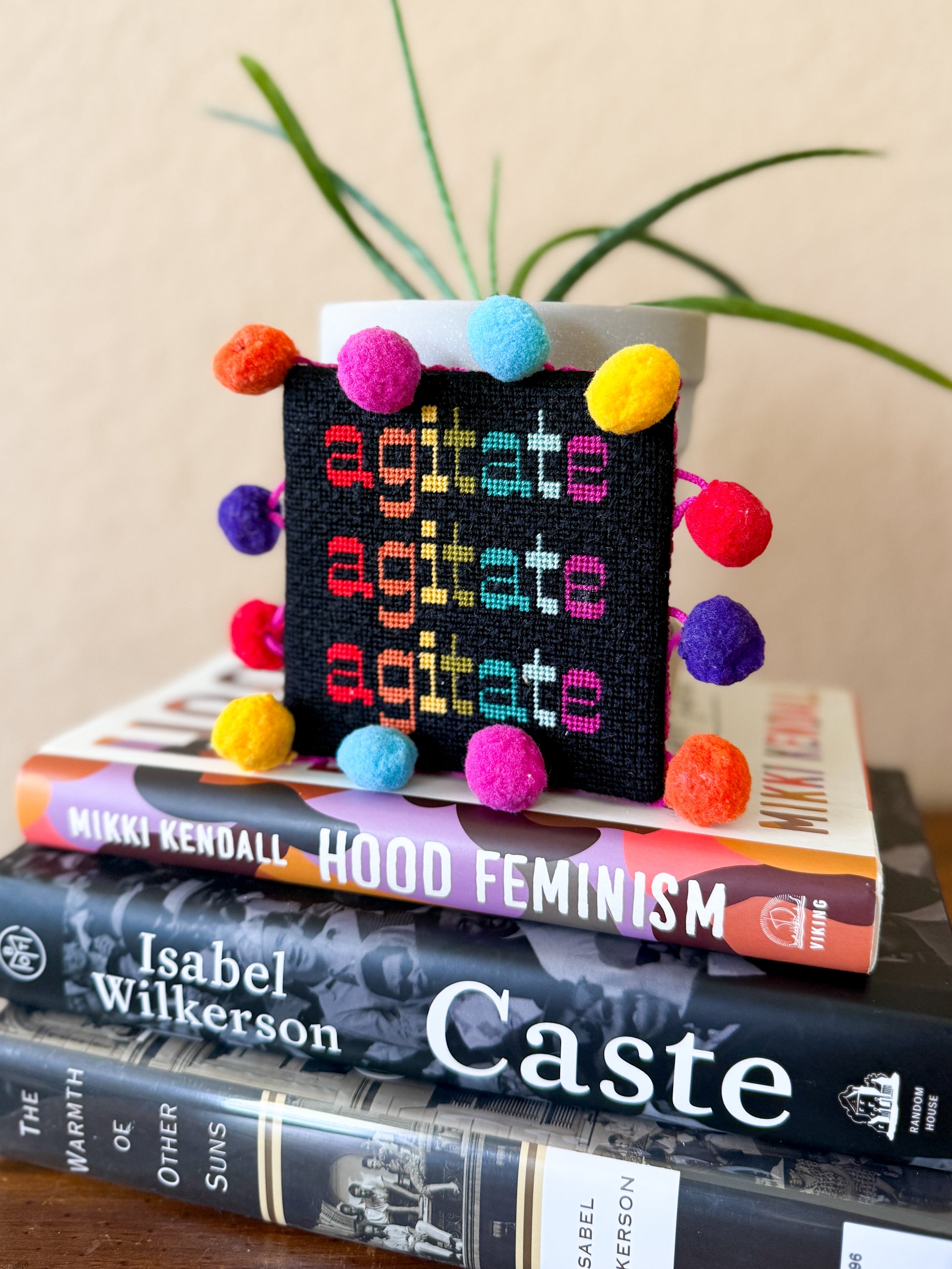 Hand-painted needlepoint canvas featuring the word “agitate” stitched three times in bright, bold colors, inspired by a quote from Frederick Douglass. Designed by Nomad & Needle.