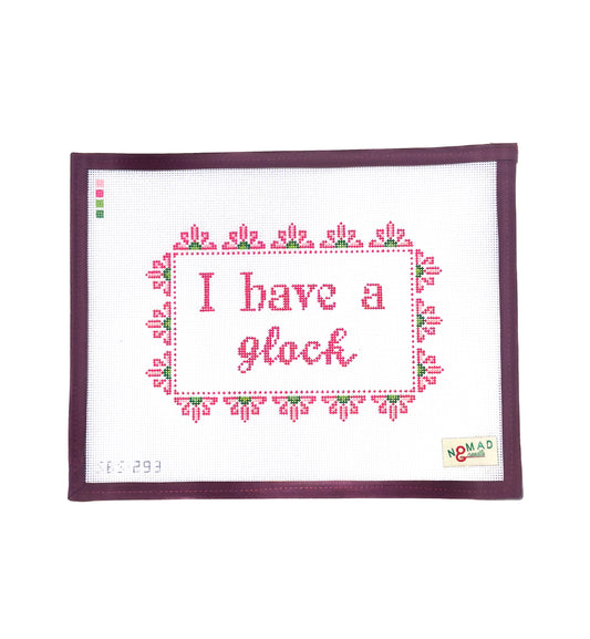 Hand-painted needlepoint canvas with the phrase “I have a glock” in pink cross-stitch lettering, framed by a floral border, designed by Nomad & Needle.
